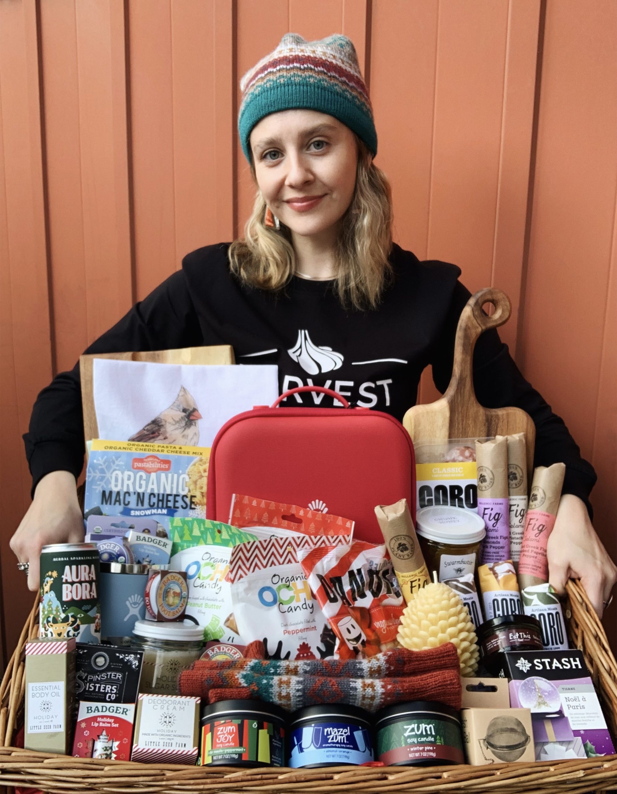 Woman with blonde hair and beanie holding a wicker basket full of holiday products like candles, candy, a red lunchbox, seasonal teas, and charcuterie products
